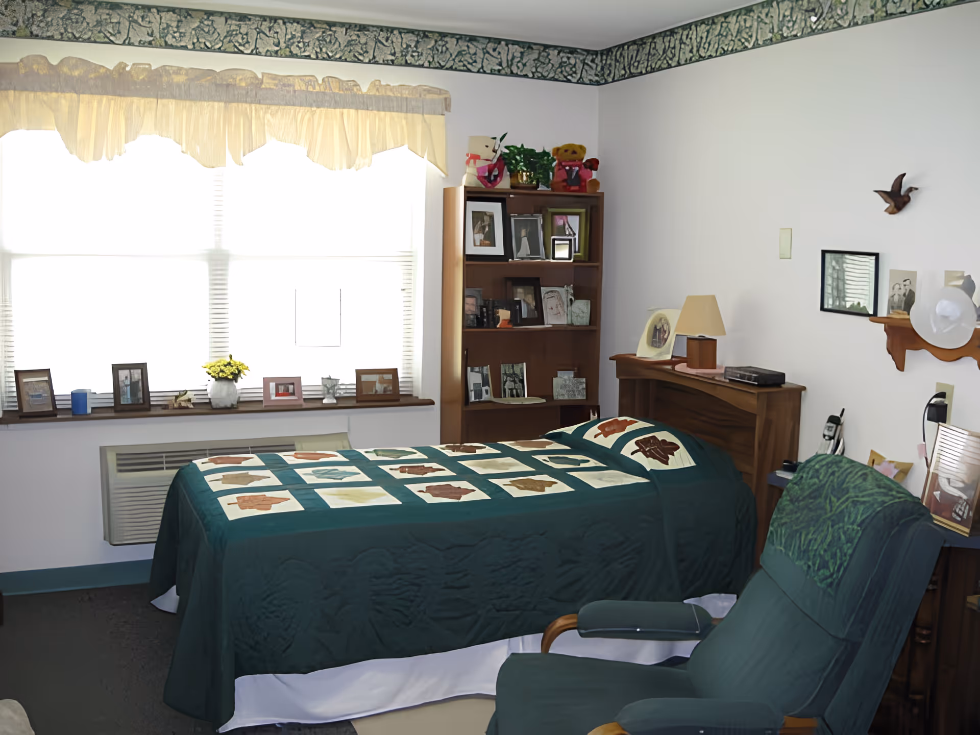 A cozy bedroom with a single bed covered in a green quilt with a patchwork design. The room has a large window with sheer curtains, a wooden bookshelf filled with framed photos and decorative items, a wooden nightstand with a lamp, and a green upholstered armchair. The walls are white with a decorative border near the ceiling and a small wooden shelf holding more framed photos and a white hat.