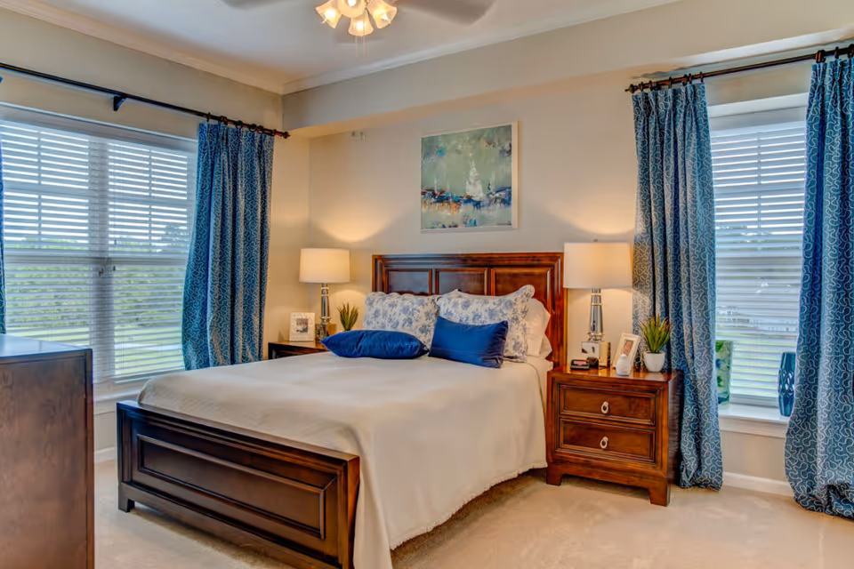 A cozy bedroom with a wooden bed frame and a beige bedspread, decorated with blue and white pillows. There are two wooden nightstands on either side of the bed, each with a lamp and decorative items. The room has two large windows with white blinds and blue patterned curtains. A painting hangs above the bed, and a ceiling fan with lights is visible.