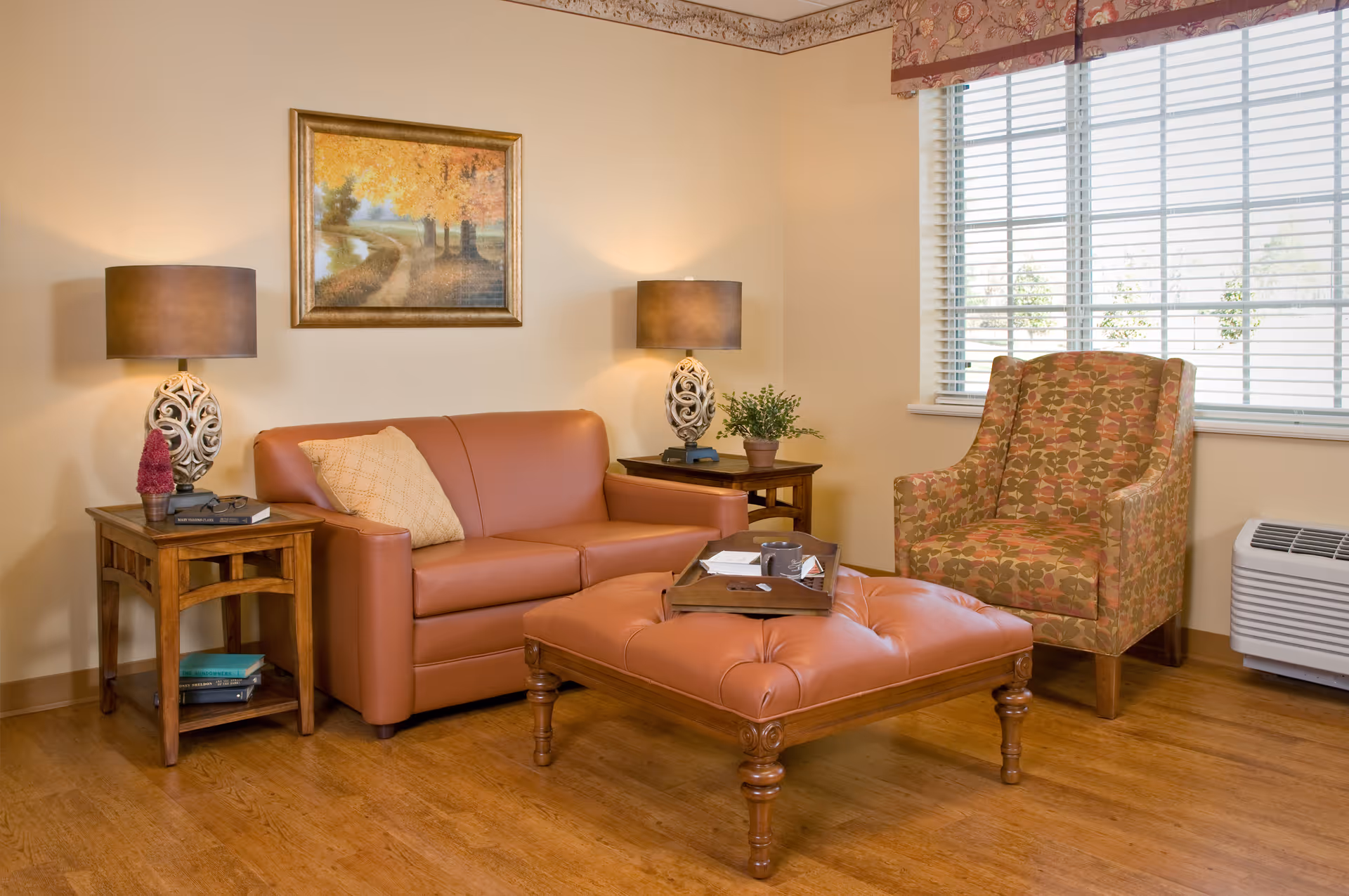 A cozy living room with a brown leather loveseat, a patterned armchair, and a large tufted ottoman serving as a coffee table. Two wooden side tables with decorative lamps and small plants flank the loveseat. A framed painting of a tree-lined path hangs on the wall above the loveseat. The room has wooden flooring and a large window with blinds and a floral valance.
