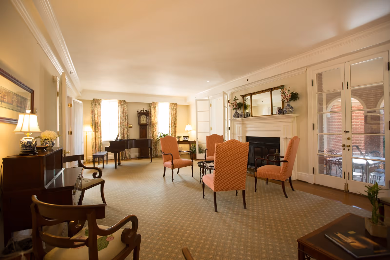 A spacious, well-lit living room with four upholstered chairs arranged around a small table near a white fireplace with a large mirror above it. The room features a grand piano, a tall grandfather clock, floral curtains on two windows, wooden furniture including chairs and cabinets, and French doors leading to an outdoor patio area.