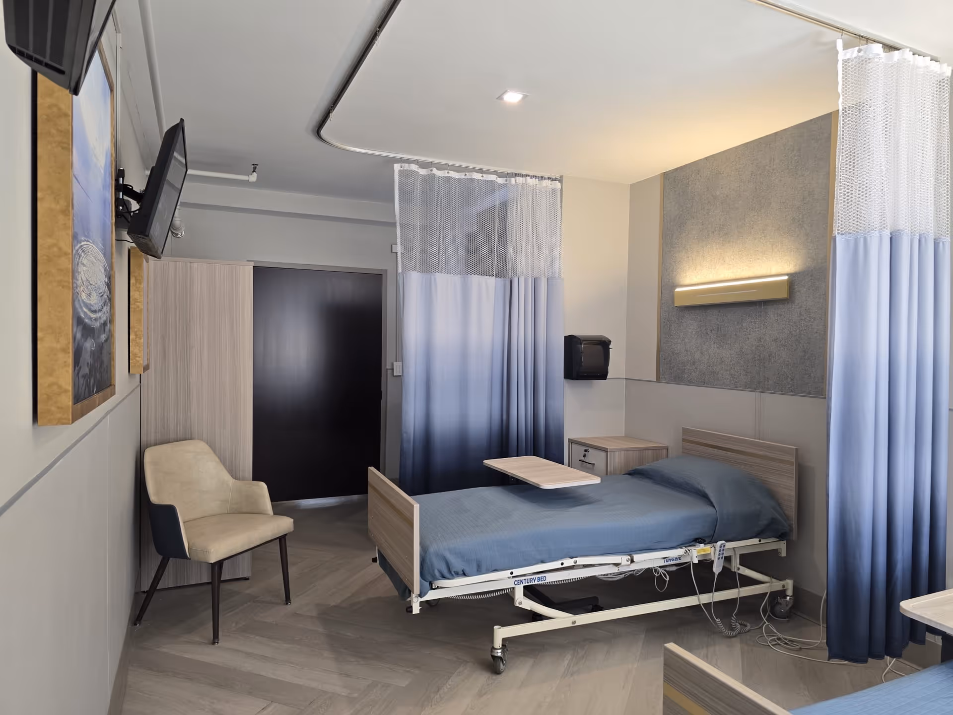 A rehabilitation or care center room with a hospital bed covered in blue bedding, a beige chair, a small wooden nightstand, a wall-mounted TV, and privacy curtains with a gradient from white to blue. The room has light-colored walls and flooring, with a modern light fixture above the bed.