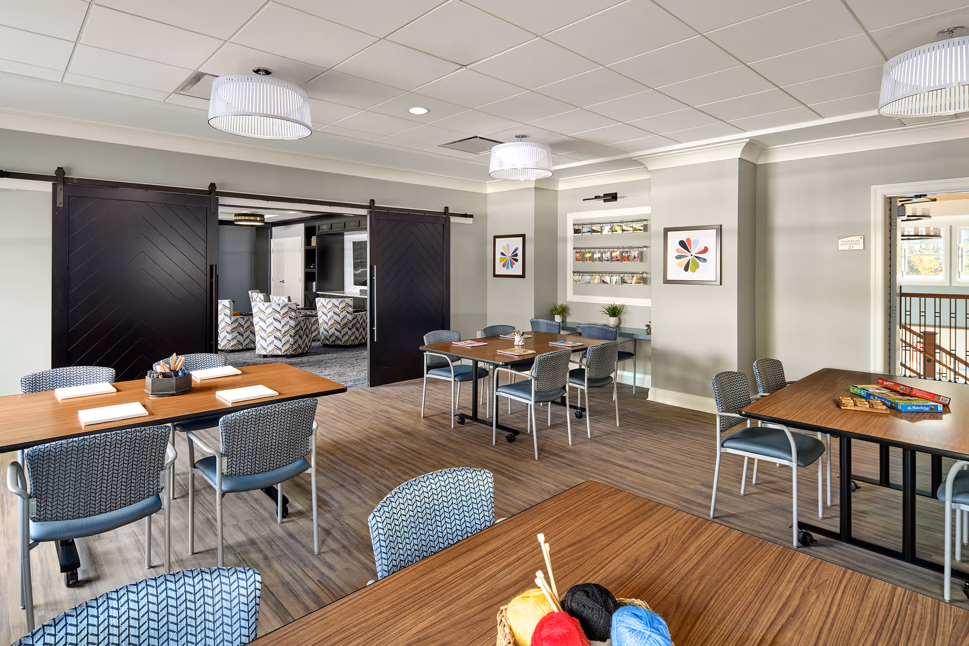 Bright multi-purpose common room with tables, chairs, craft supplies and sliding barn doors opening to a lounge area.