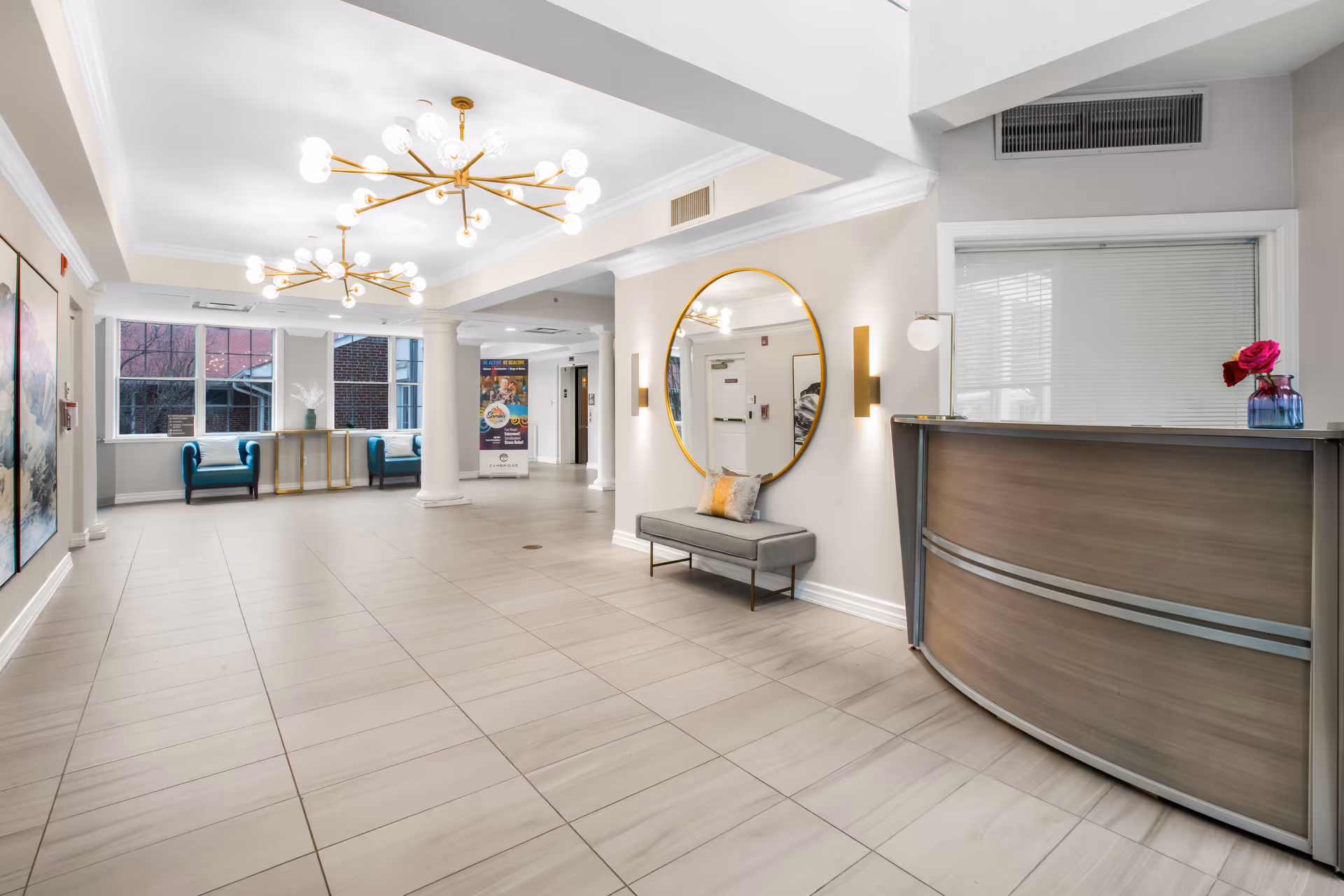 A spacious and well-lit senior living facility lobby with modern decor. The area features large beige floor tiles, two contemporary gold chandeliers with globe lights, a round gold-framed mirror above a gray bench with pillows, and a curved reception desk with a small vase holding pink flowers. There are two blue armchairs and a console table near large windows letting in natural light. The walls are light-colored, and there are columns and artwork visible in the space.