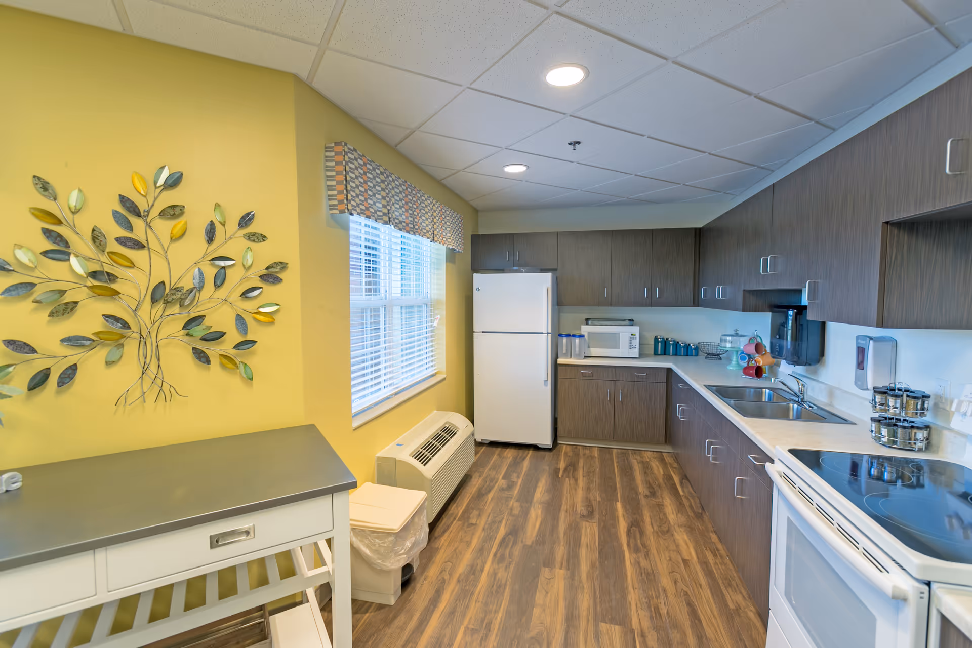 Well-lit communal kitchen with a yellow accent wall featuring metal leaf art, white refrigerator and appliances, sink, stove, and brown cabinets.