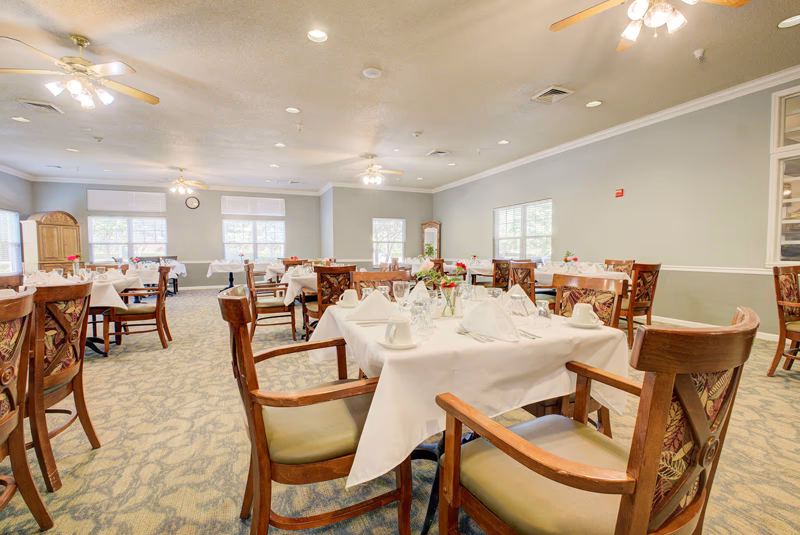 A bright and spacious dining room with multiple tables covered in white tablecloths, each set with napkins, cups, glasses, and silverware. The room features wooden chairs with patterned cushions, large windows letting in natural light, ceiling fans with lights, and a carpeted floor.