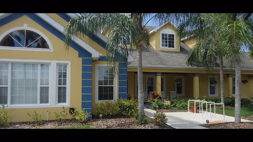 Front entrance of a yellow and blue single-story building with palm trees, a covered porch with rocking chairs, walkway and landscaping.
