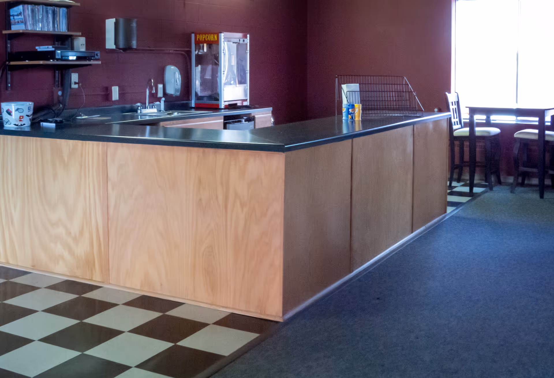 Interior view of a kitchen area with a long wooden counter topped with a black countertop. Behind the counter, there is a sink, a popcorn machine, and shelves with electronic devices. To the right, there is a dining area with a table and chairs near a window letting in natural light. The floor has a checkered pattern near the counter and carpet in the dining area.