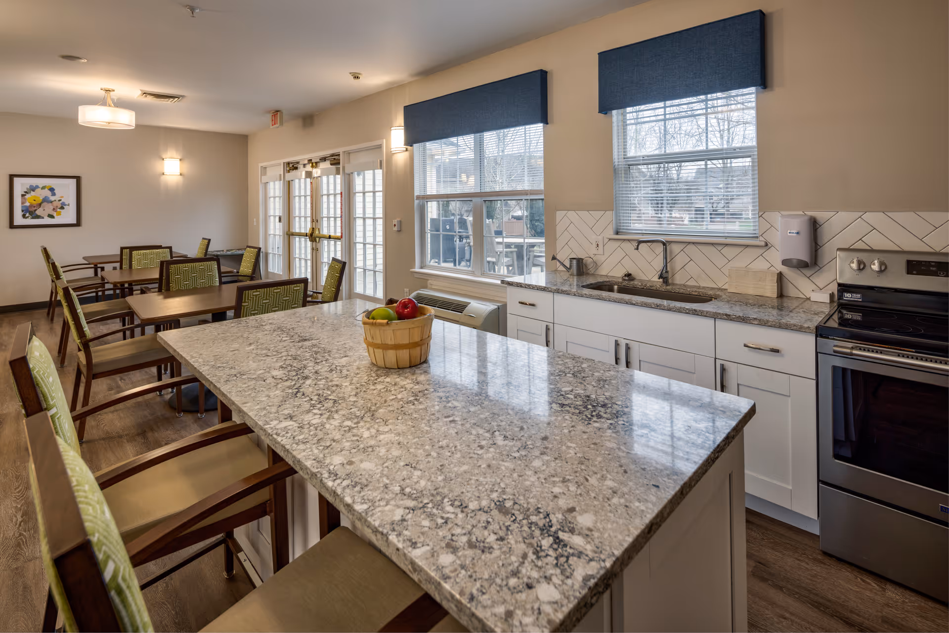 A bright and clean kitchen and dining area in a senior living facility. The kitchen features a large granite island with chairs, a basket of apples on the island, white cabinets, a stainless steel oven, and a sink under a window with blue valances. The dining area has several wooden tables and chairs with green patterned cushions, and there is a framed floral artwork on the wall. Large windows and glass doors allow natural light to fill the space.