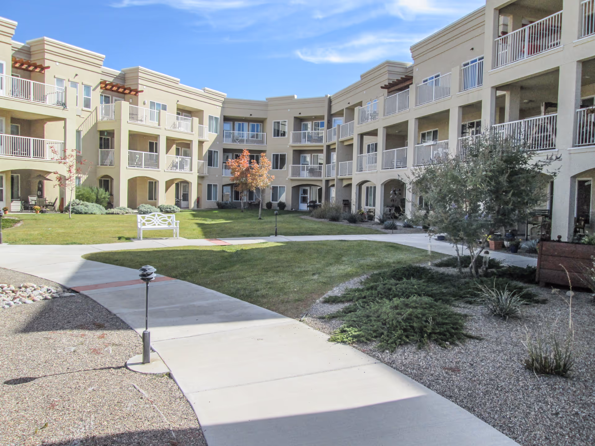 Outdoor courtyard area of a senior living facility with a well-maintained lawn, small trees, shrubs, and a white bench. The courtyard is surrounded by a three-story beige building with balconies and large windows under a blue sky with some clouds.