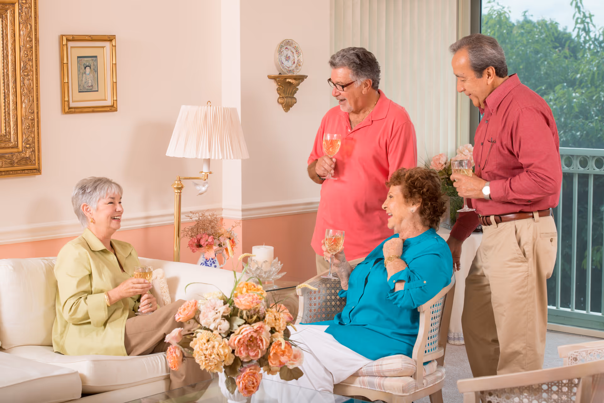Four elderly adults socializing and enjoying drinks in a well-lit living room with floral decorations, a lamp, and framed artwork on the walls. Two women are seated, one on a white couch and the other on a chair, while two men stand nearby holding glasses. A large window with vertical blinds and greenery outside is visible in the background.