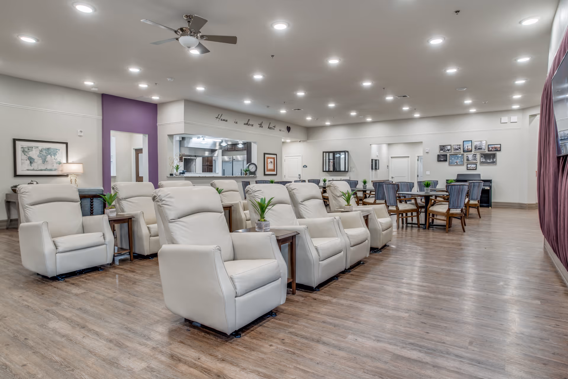 A spacious and well-lit common area in a senior living facility featuring multiple beige recliner chairs arranged in rows with small tables and potted plants between them. Behind the chairs are several round tables with striped upholstered chairs. The room has wood flooring, recessed ceiling lights, a ceiling fan, and a wall with the phrase 'Home is where the heart is' written above an open kitchen area. There are framed pictures on the walls and a large TV mounted on the right wall with curtains beside it.