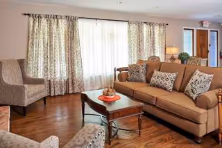 A cozy living room with a brown sofa adorned with patterned throw pillows, two upholstered armchairs, a wooden coffee table with decorative items, hardwood flooring, and large windows covered with patterned curtains allowing natural light to enter.