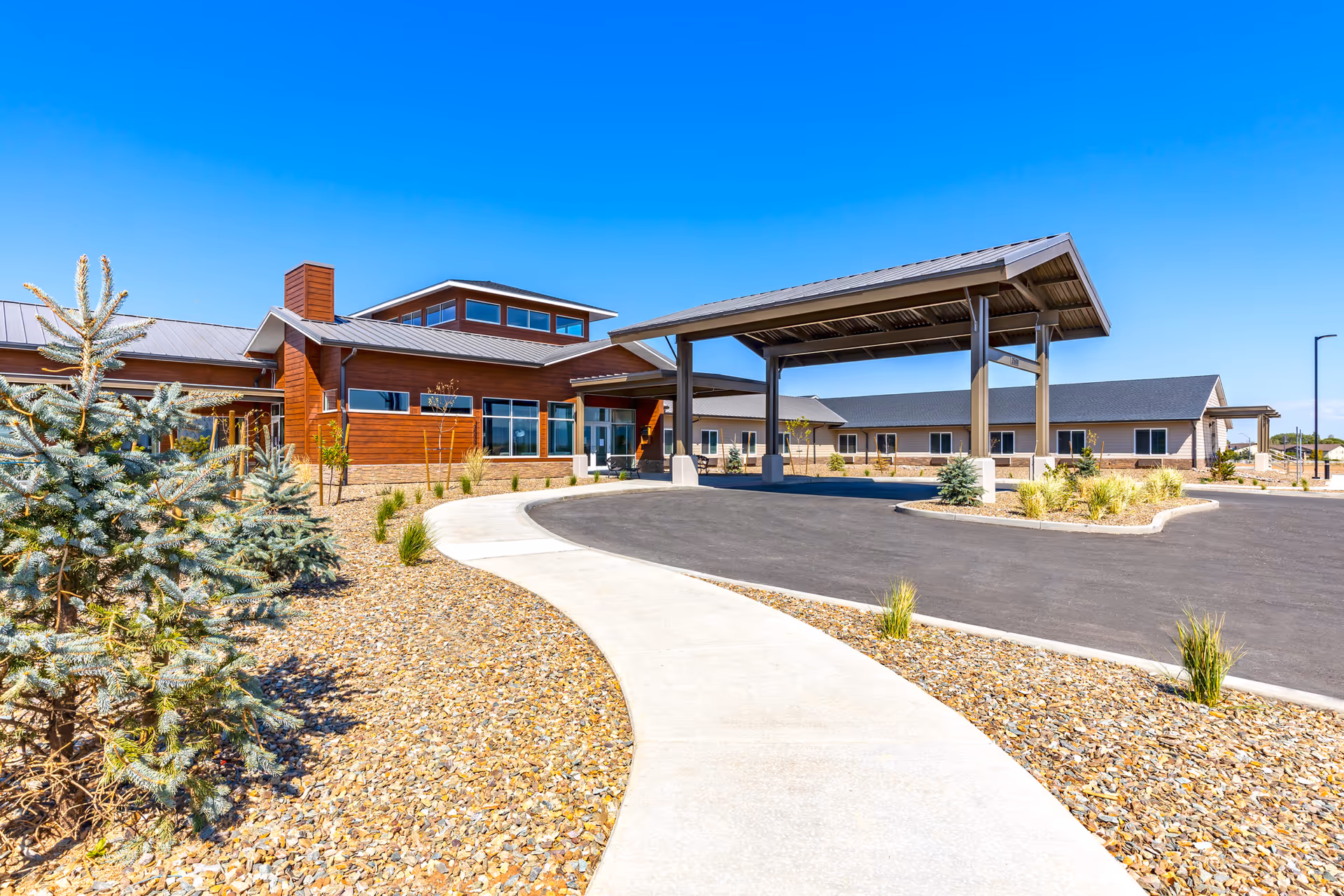 Front entrance of a single-story senior living facility with a covered drop-off porte-cochere, curved walkway, and drought-tolerant landscaping under a clear blue sky.