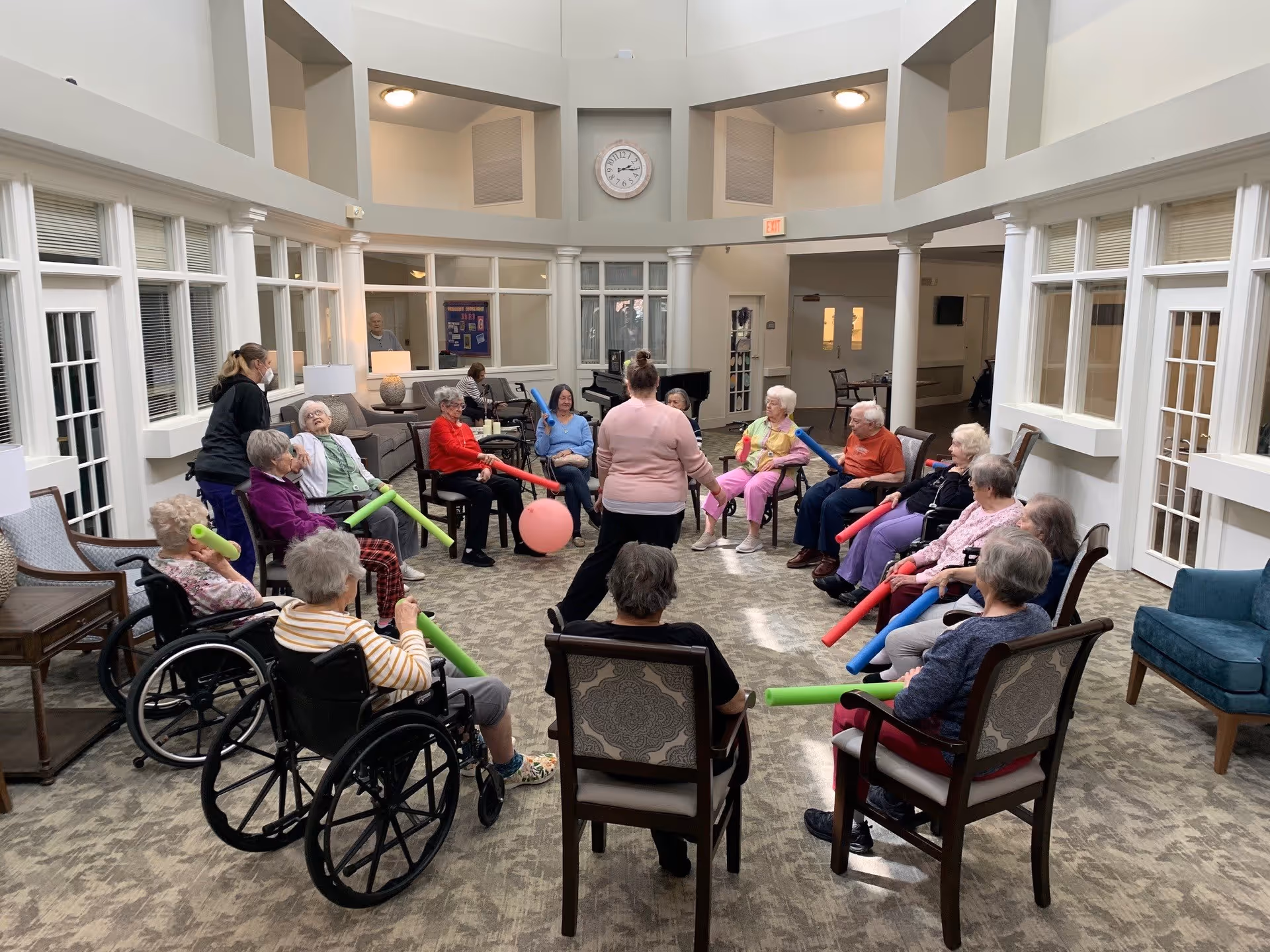 A group of elderly individuals seated in a circle in a spacious, well-lit common area with large windows and high ceilings. They are participating in a group activity using colorful foam sticks and a pink ball, led by a standing instructor. Some participants are in wheelchairs, and the room has a carpeted floor and comfortable chairs.