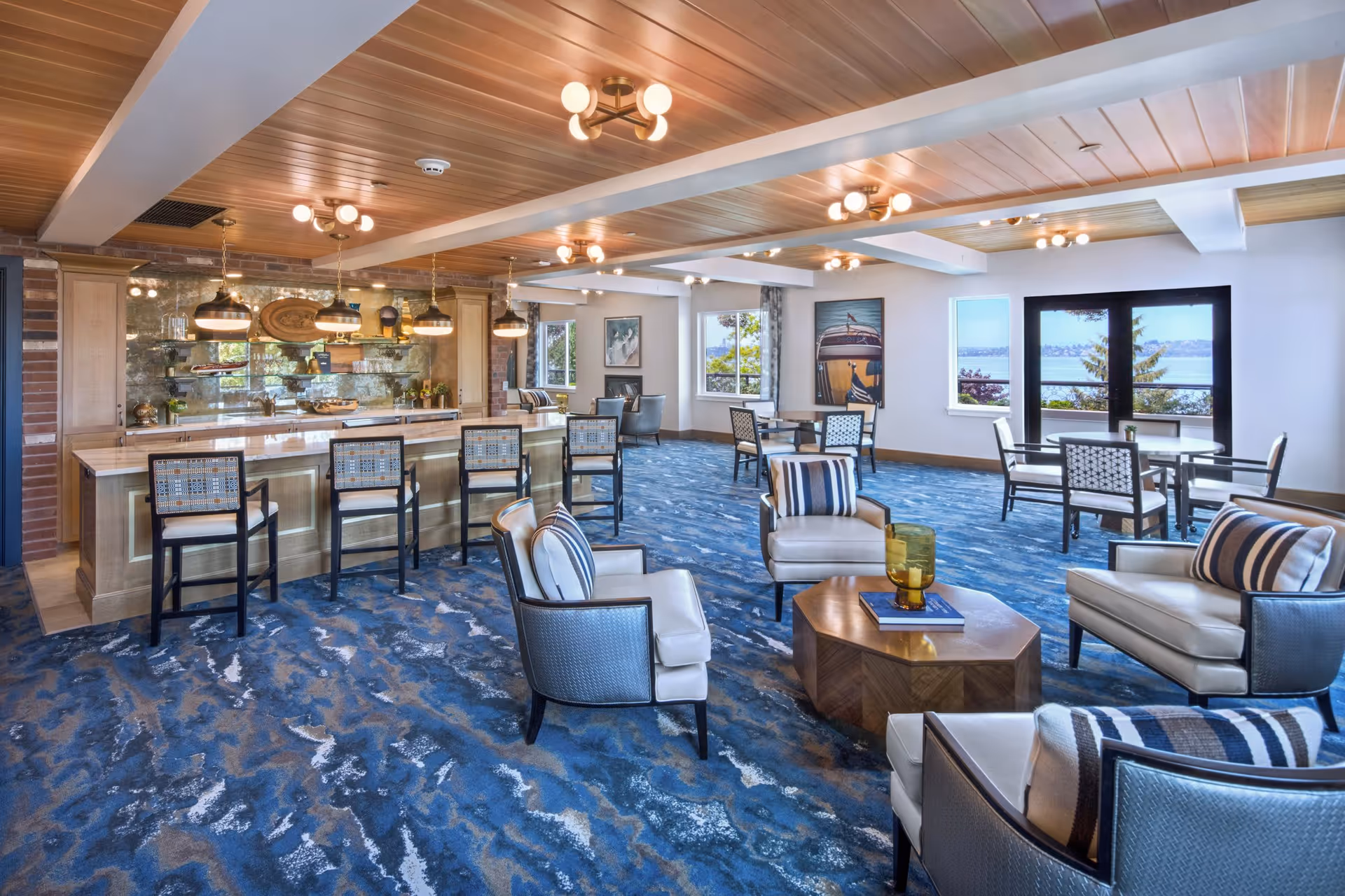A spacious and well-lit common area with a blue patterned carpet, several comfortable armchairs with striped pillows arranged around a wooden coffee table, a bar counter with high chairs, pendant lights hanging above the bar, and multiple tables and chairs near large windows offering a view of water and trees outside.