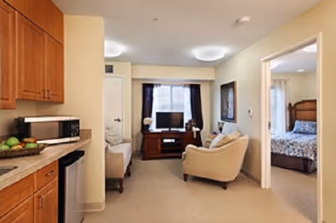 Assisted-living suite interior with a kitchenette at left, a seating area with a TV in the center, and a bedroom visible through an open doorway.