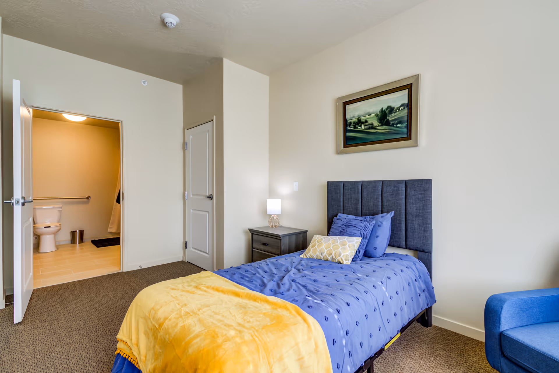 A senior living facility bedroom with a single bed covered in a blue bedspread and a yellow blanket. There is a nightstand with a lamp next to the bed, a framed landscape painting on the wall above the bed, and a blue armchair partially visible on the right. An open door reveals an adjacent bathroom with a toilet and grab bar.