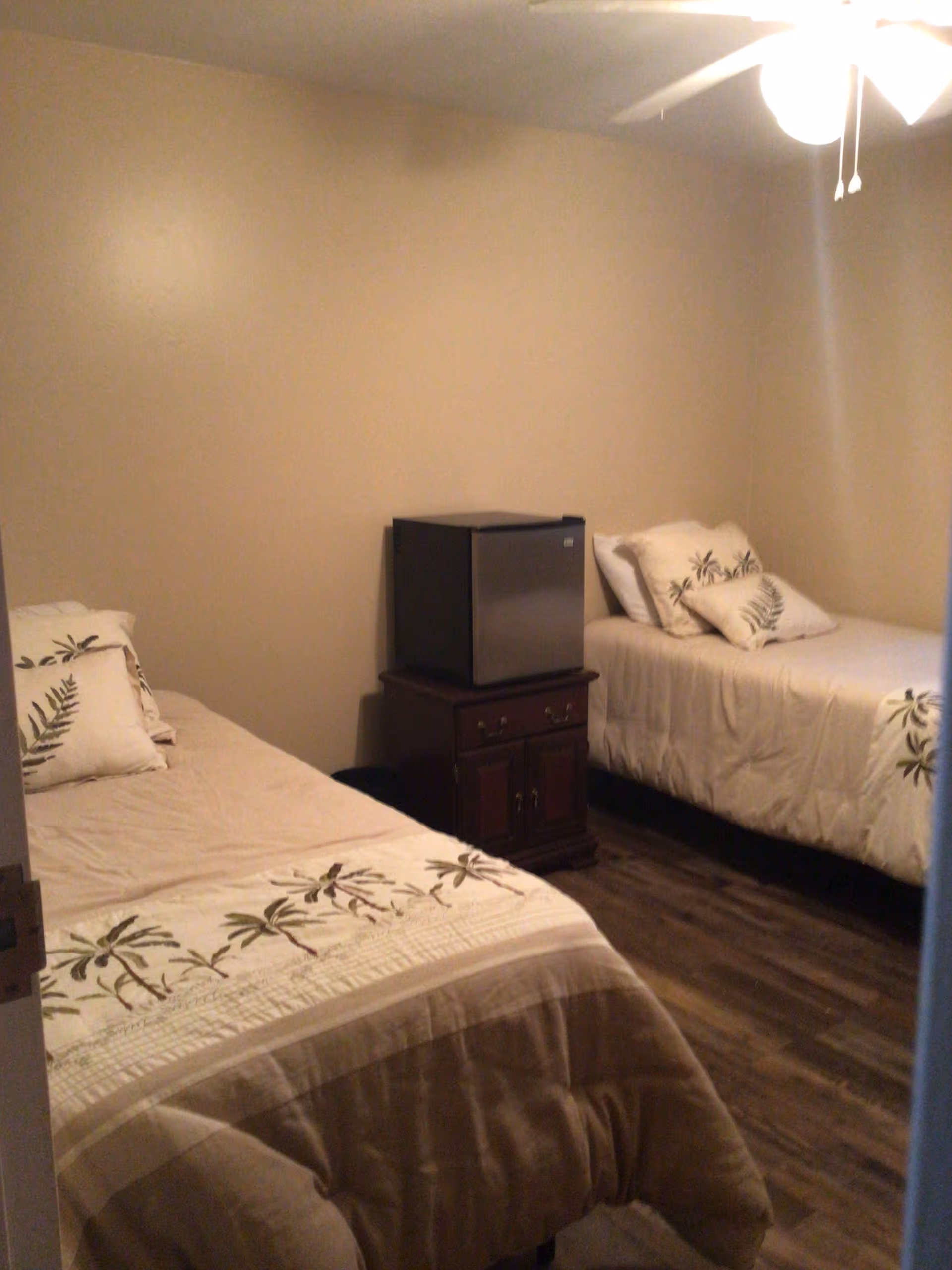 A small bedroom with two single beds, each with beige bedding and decorative pillows featuring leaf patterns. Between the beds is a wooden nightstand with a small black mini fridge on top. The room has beige walls and wood flooring, with a ceiling fan and light fixture visible at the top right.
