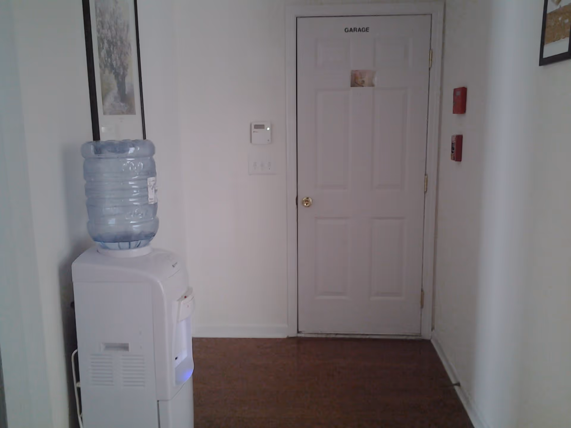 Indoor hallway area with a white water cooler on the left side, a closed door labeled 'GARAGE' straight ahead, a thermostat on the wall, and two red fire alarm devices mounted on the right wall.