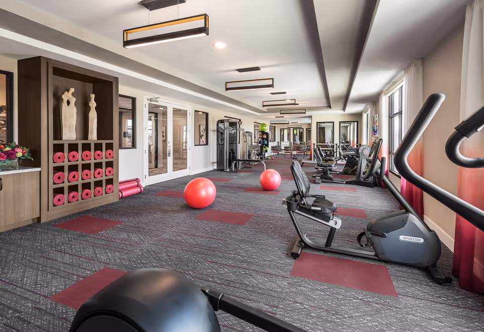 A spacious fitness room with exercise equipment including stationary bikes, weight machines, and two large pink exercise balls on a carpeted floor with red and gray patterns. The room has large windows with red curtains allowing natural light to enter. On the left side, there is a wooden shelving unit holding neatly rolled pink yoga mats and two decorative sculptures. Modern rectangular ceiling lights illuminate the room.
