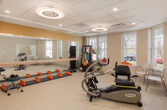 Bright fitness room with a NuStep recumbent trainer, exercise mats with orange step platforms, weight rack and a mirrored wall.