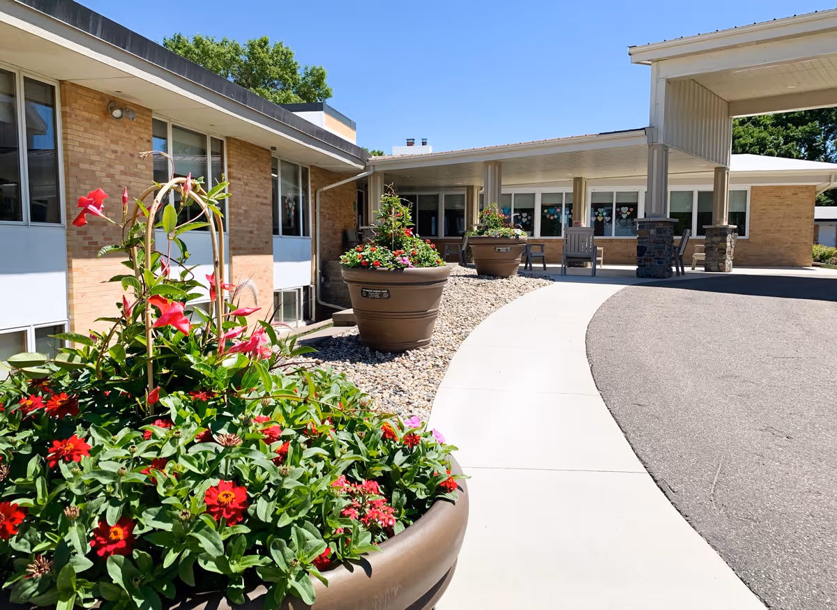 Outdoor view of Glenwood Retirement Village showing a curved driveway and sidewalk leading to the building entrance. Large flower pots with vibrant red and pink flowers line the sidewalk. The building has a brick exterior with large windows and a covered entrance area with chairs.
