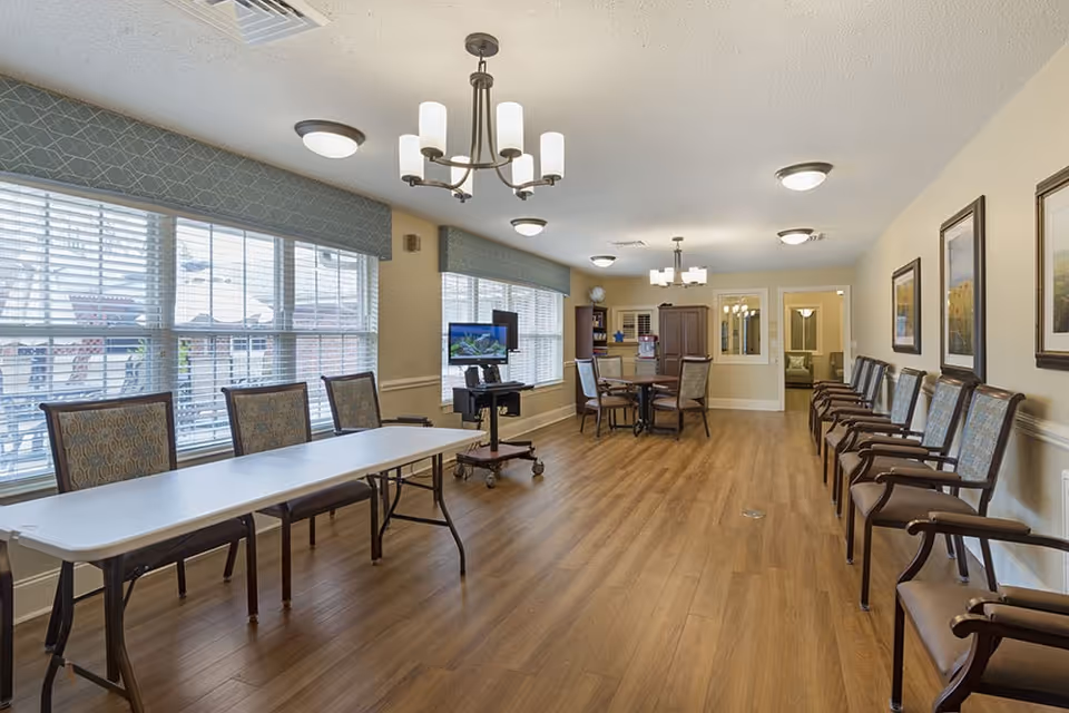 A spacious, well-lit room with wooden flooring and large windows covered with blinds. The room features a long white folding table with chairs on one side, a round table with chairs in the background, and a row of armchairs lined up against the opposite wall. The walls are decorated with framed artwork, and there are multiple ceiling light fixtures including chandeliers. A TV on a stand is positioned near the windows.