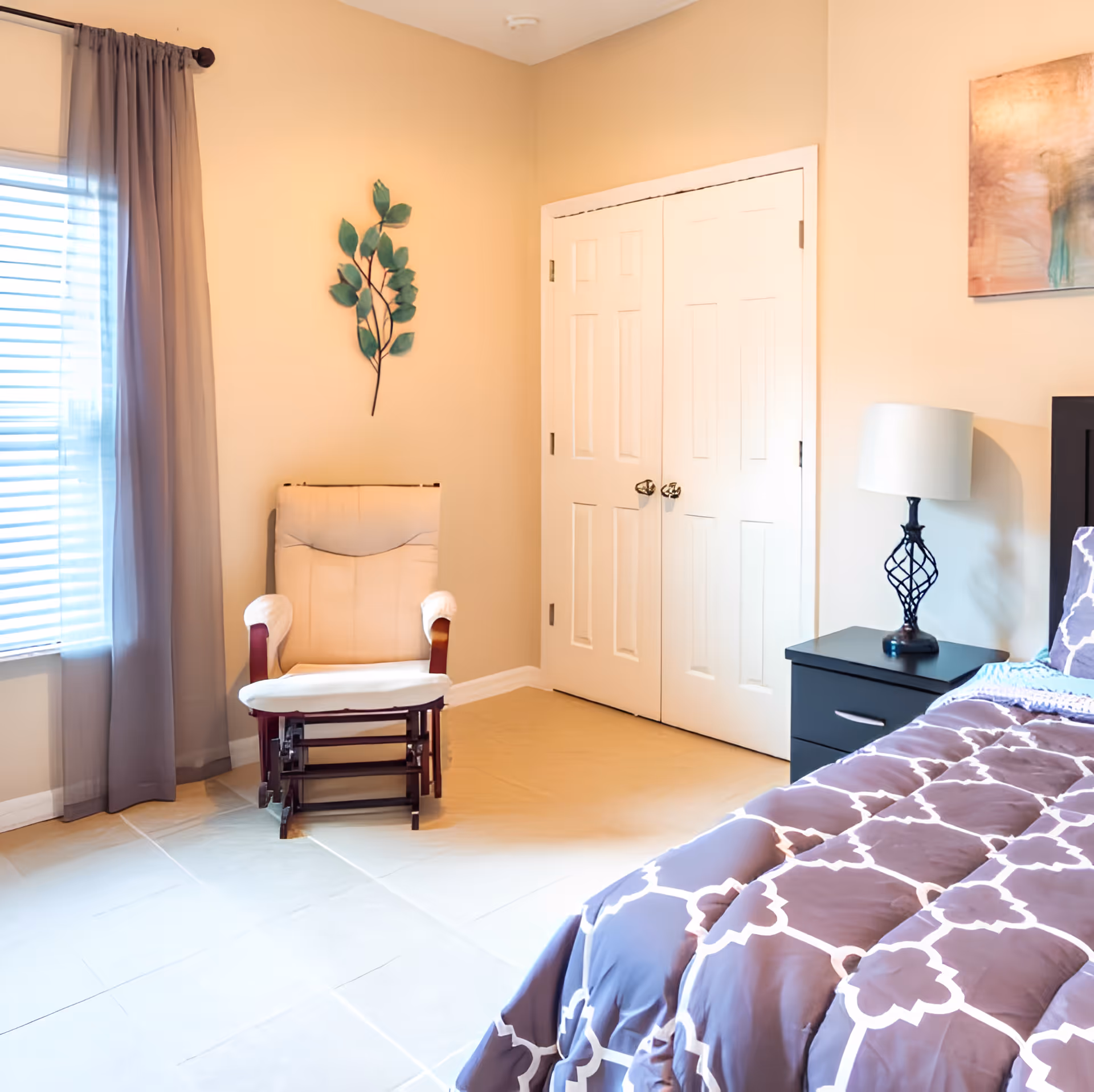 A bright bedroom with a patterned bedspread, nightstand and lamp, a cushioned rocking chair, closet doors, and a curtained window.