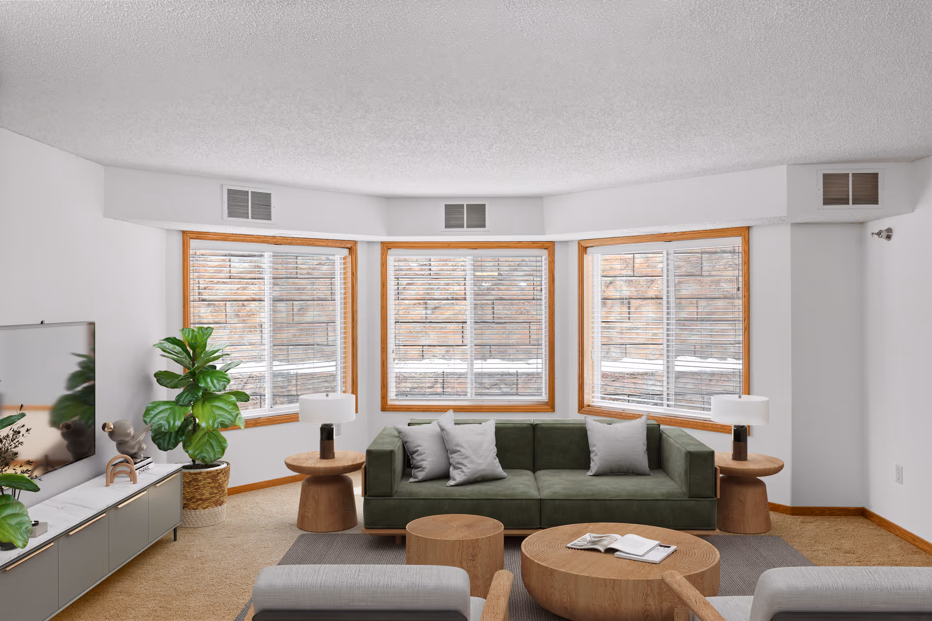 A bright living room with three large windows featuring wooden frames and white blinds. The room has a green sofa with three gray cushions, two wooden side tables with white lamps, a round wooden coffee table with an open book, and a gray armchair. There is a TV on a low cabinet with decorative plants and a sculpture, and a large green potted plant in the corner. The walls are white, and the floor is carpeted in beige.