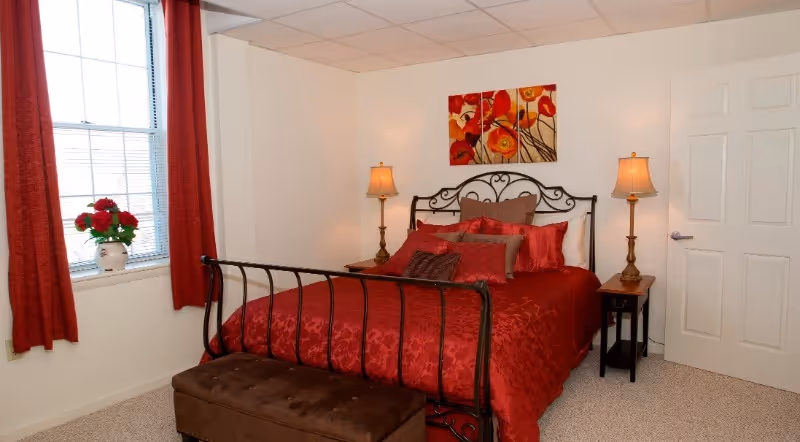 A bedroom with a black metal bed frame and red bedding, including multiple pillows. There are two bedside tables with matching lamps on each side of the bed. A brown cushioned bench is at the foot of the bed. A window with red curtains and a vase of red flowers is on the left wall. A three-panel floral painting hangs above the bed on a white wall.