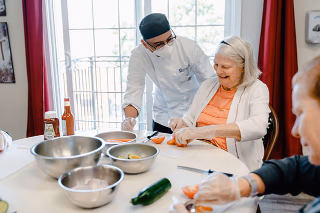 A chef wearing a white uniform and black hat assists an elderly woman seated at a table as she cuts a tomato. Several metal bowls with ingredients and a cucumber are on the table. Another elderly person is partially visible in the foreground, also engaged in food preparation.