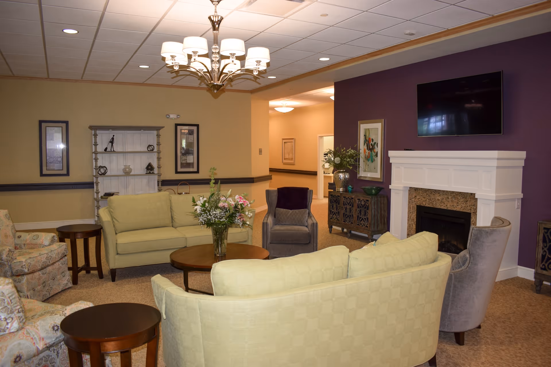 A cozy living room area in a senior living facility with light green sofas, patterned armchairs, a round wooden coffee table with a vase of flowers, a white fireplace with a mounted flat-screen TV above it, and a purple accent wall. There are framed artworks on the walls and a chandelier hanging from the ceiling.