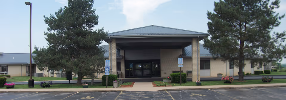 Front exterior view of Lawrence County Residential Care Center building with a covered entrance, two large pine trees, flower pots, and several handicap parking spaces in front.