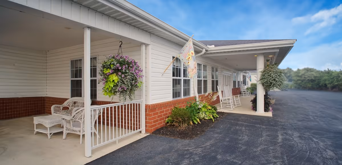 Covered porch area of a senior living facility with white wicker chairs and a hanging flower basket. The building has white siding with a red brick lower section and several windows. There are also rocking chairs along the porch and a flag hanging near the entrance. The paved driveway extends alongside the building with greenery in the background under a blue sky.