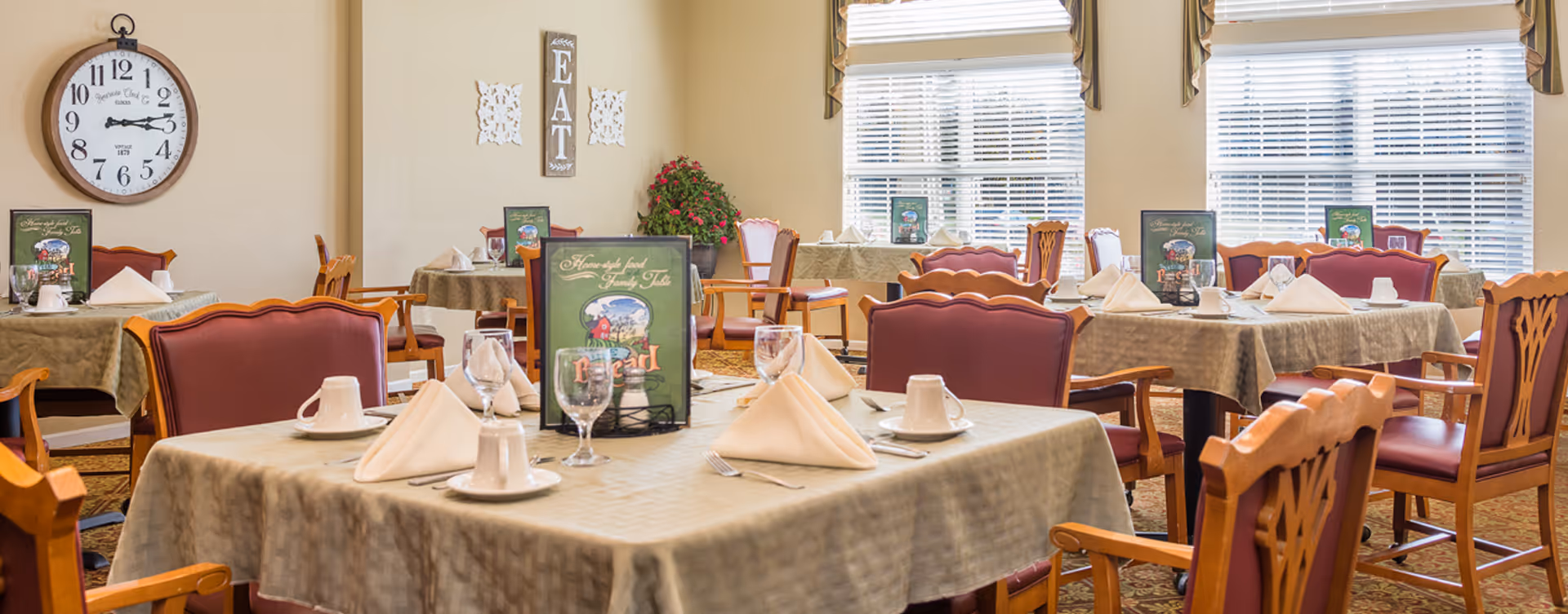 A dining room with several tables covered in beige tablecloths, each set with folded napkins, cups, glasses, and menus. The chairs have wooden frames with red cushioned seats and backs. Large windows with blinds allow natural light to fill the room. A wall clock and a decorative sign that says 'EAT' are visible on the wall.
