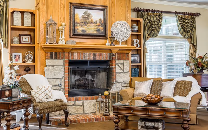Cozy living room with a stone and brick fireplace as the focal point, flanked by wooden built-in shelves displaying decorative items and framed pictures. A patterned armchair with a white throw and pillow is on the left, and a beige sofa with white and brown pillows is on the right. A wooden coffee table with a glass top and a decorative bowl sits in front of the sofa. Large windows with patterned curtains allow natural light to fill the room.