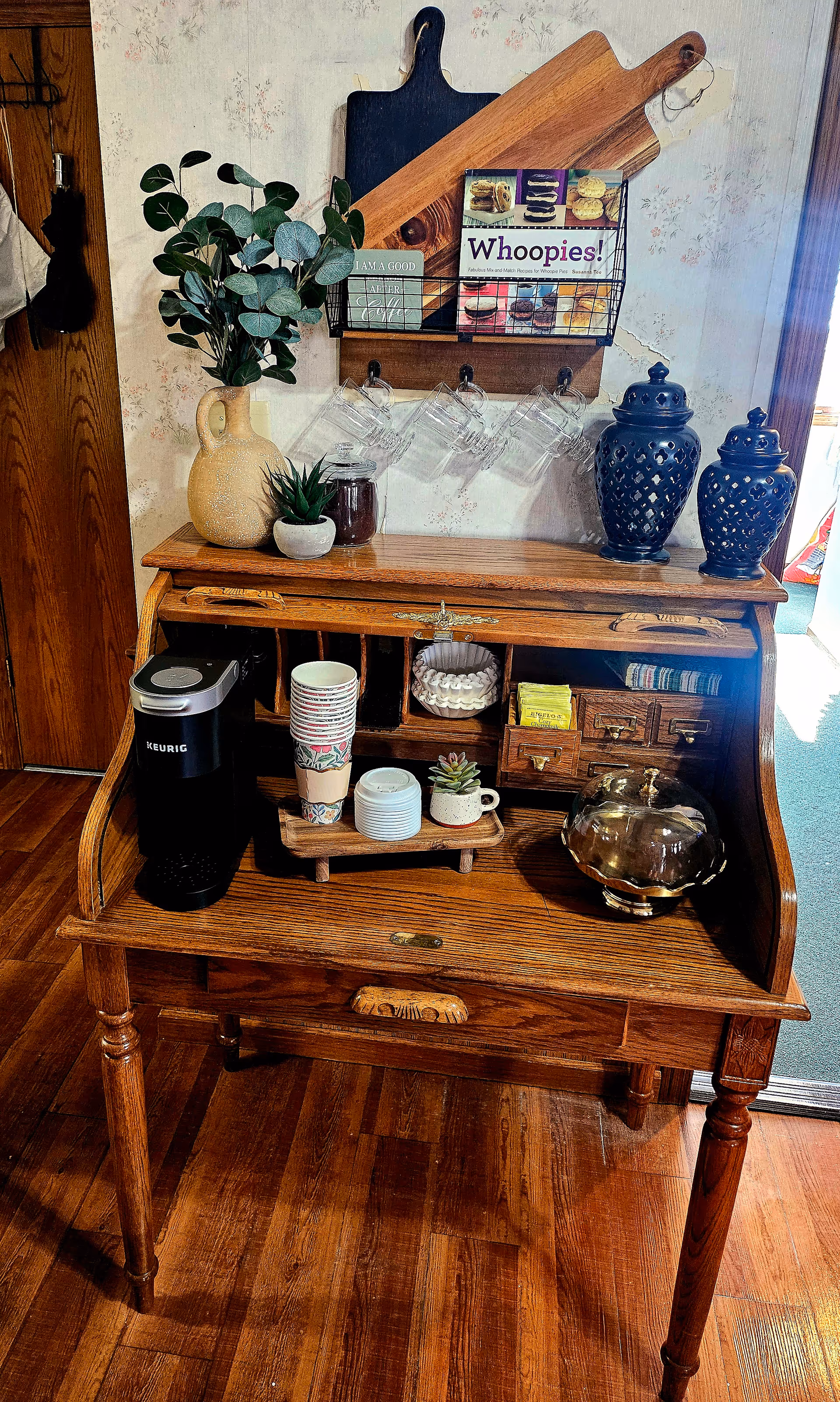 A wooden roll-top desk set up as a coffee station with a Keurig coffee maker, stacked disposable cups, small potted plants, a silver bowl, and various small drawers. Above the desk, there are two wooden cutting boards, a wire rack holding a cookbook titled 'Whoopies!', and four hanging glass mugs. The background features a floral wallpaper and a wooden door to the left.