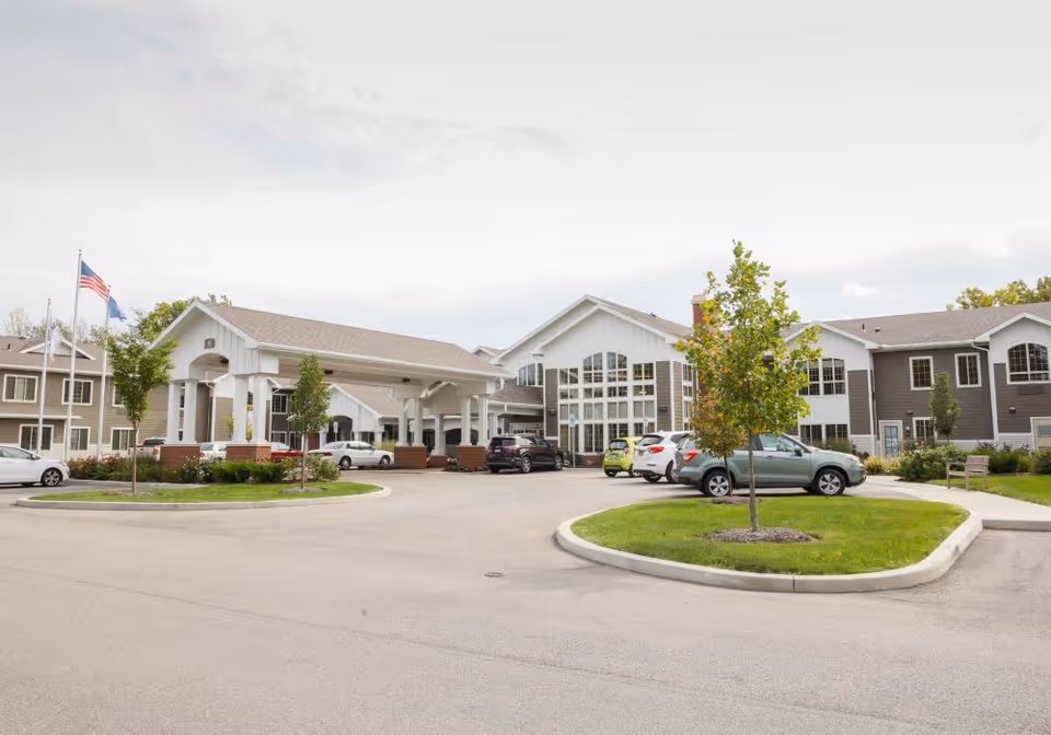 Front entrance of a senior living facility with a covered porte-cochere, landscaped islands, parked cars, and flags.