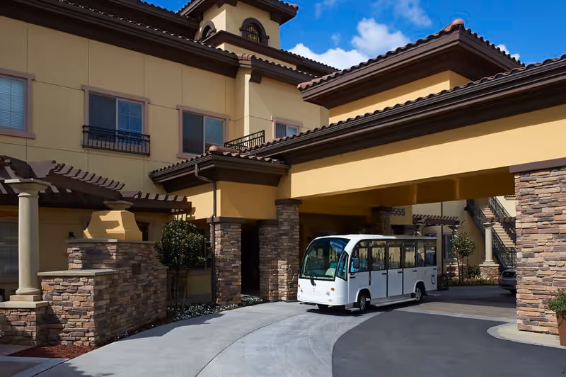 Exterior view of a senior living facility with a covered driveway entrance. A white electric shuttle vehicle is parked under the covered area. The building features stone and stucco walls with a tiled roof and multiple windows. There are small trees and landscaping around the entrance.