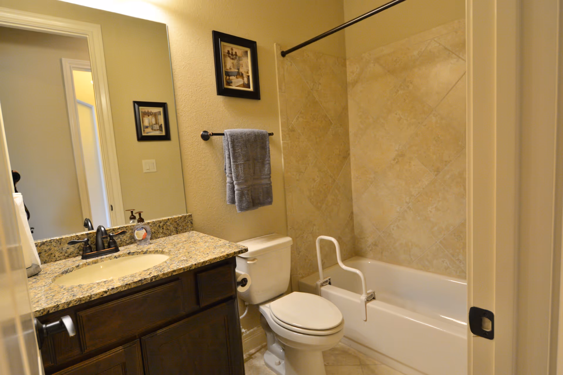 A bathroom with a granite countertop sink, a toilet, and a bathtub with beige tiled walls. There is a towel rack with a gray towel above the toilet and two framed pictures on the wall. A white safety grab bar is attached to the bathtub.