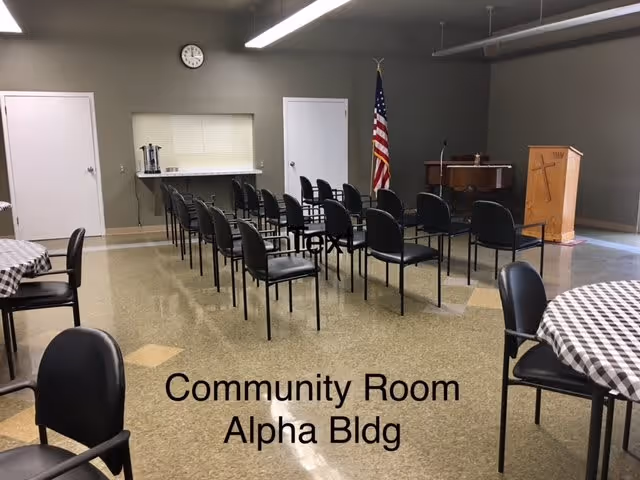 Interior view of a community room in the Alpha Building with rows of black chairs facing a wooden podium and an American flag. There are round tables with checkered tablecloths and chairs around them. A clock is mounted on the wall above two closed doors.