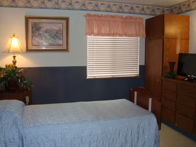 A small bedroom in a senior living facility with a single bed covered in a light blue quilt. There is a wooden nightstand with a lamp and a plant on it to the left of the bed. A framed picture hangs on the wall above the nightstand. The window has closed blinds and a pink valance. On the right side of the room, there is a wooden wardrobe and a dresser with a small TV on top.