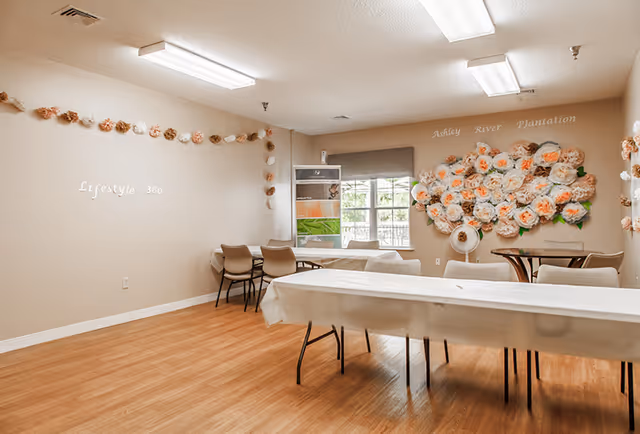 A bright room with wooden flooring and beige walls decorated with paper flowers and garlands. There are several tables covered with white tablecloths and chairs arranged around them. On one wall, there is a large floral arrangement with the words 'Ashley River Plantation' above it. Another wall has the words 'Lifestyle 360'. A window with a gray blind lets in natural light.