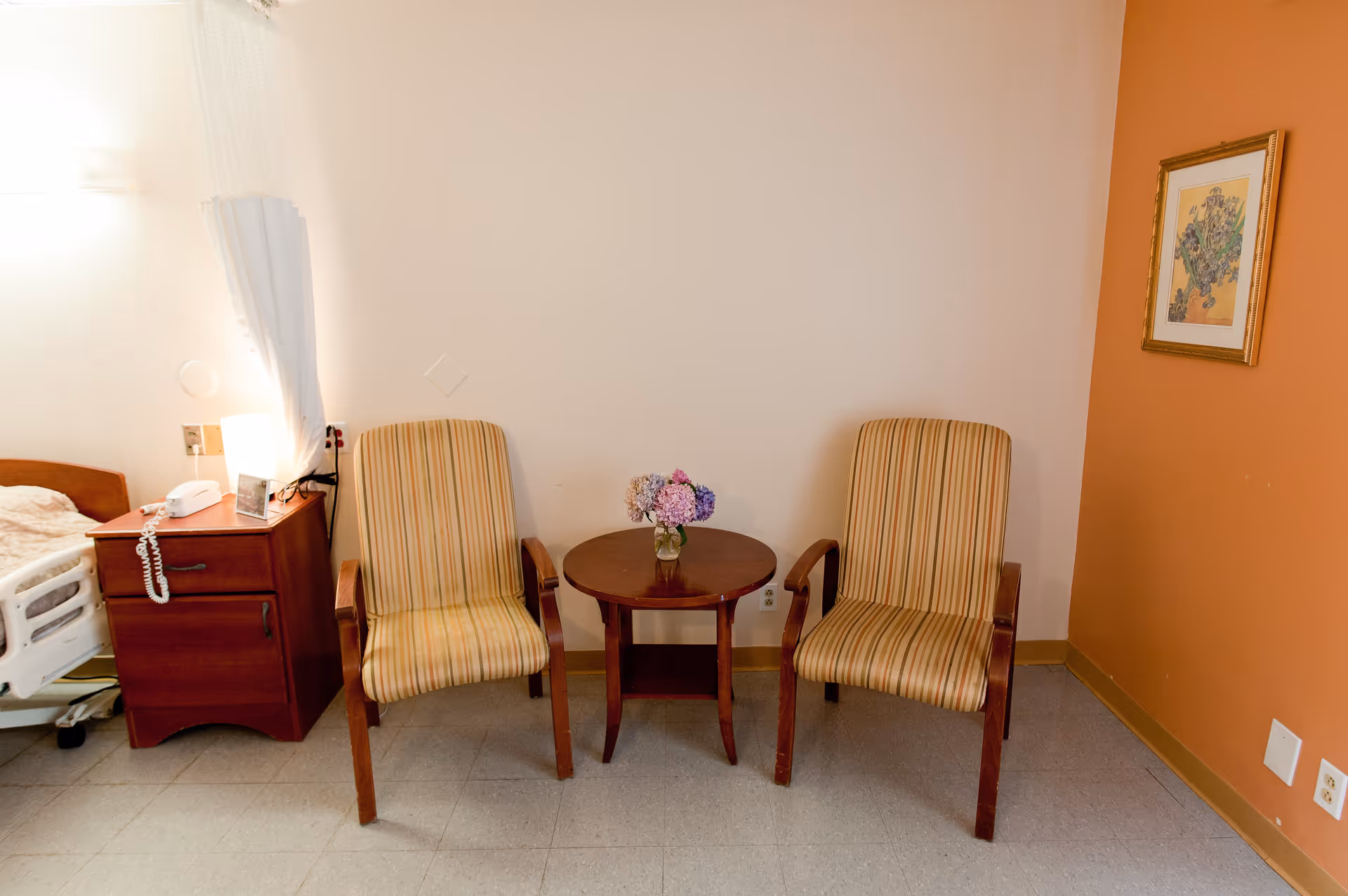 Two striped armchairs flank a small round table holding a vase of flowers in a senior living room with a bedside table and a partially visible hospital bed.