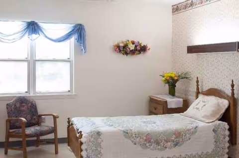 A cozy bedroom in a senior care facility with a bed, chair, nightstand, and flowers.