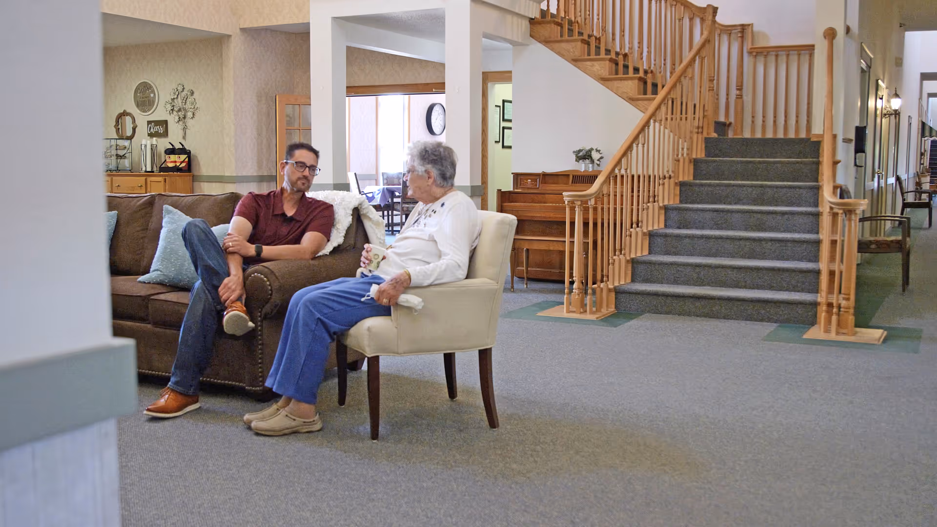 An elderly woman and a man sitting and talking in a living room area of a senior living facility. The woman is seated in a beige armchair holding a cup, while the man is sitting on a brown couch. Behind them is a wooden staircase with carpeted steps, a piano, and a hallway leading to other rooms.