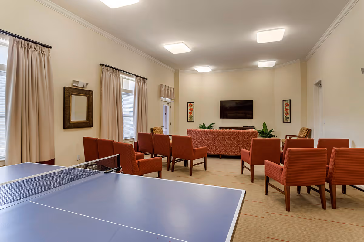 A community room with a blue ping pong table in the foreground and several red upholstered chairs arranged facing a wall-mounted flat screen TV. The room has beige walls, large windows with beige curtains, framed floral artwork, and ceiling lights.