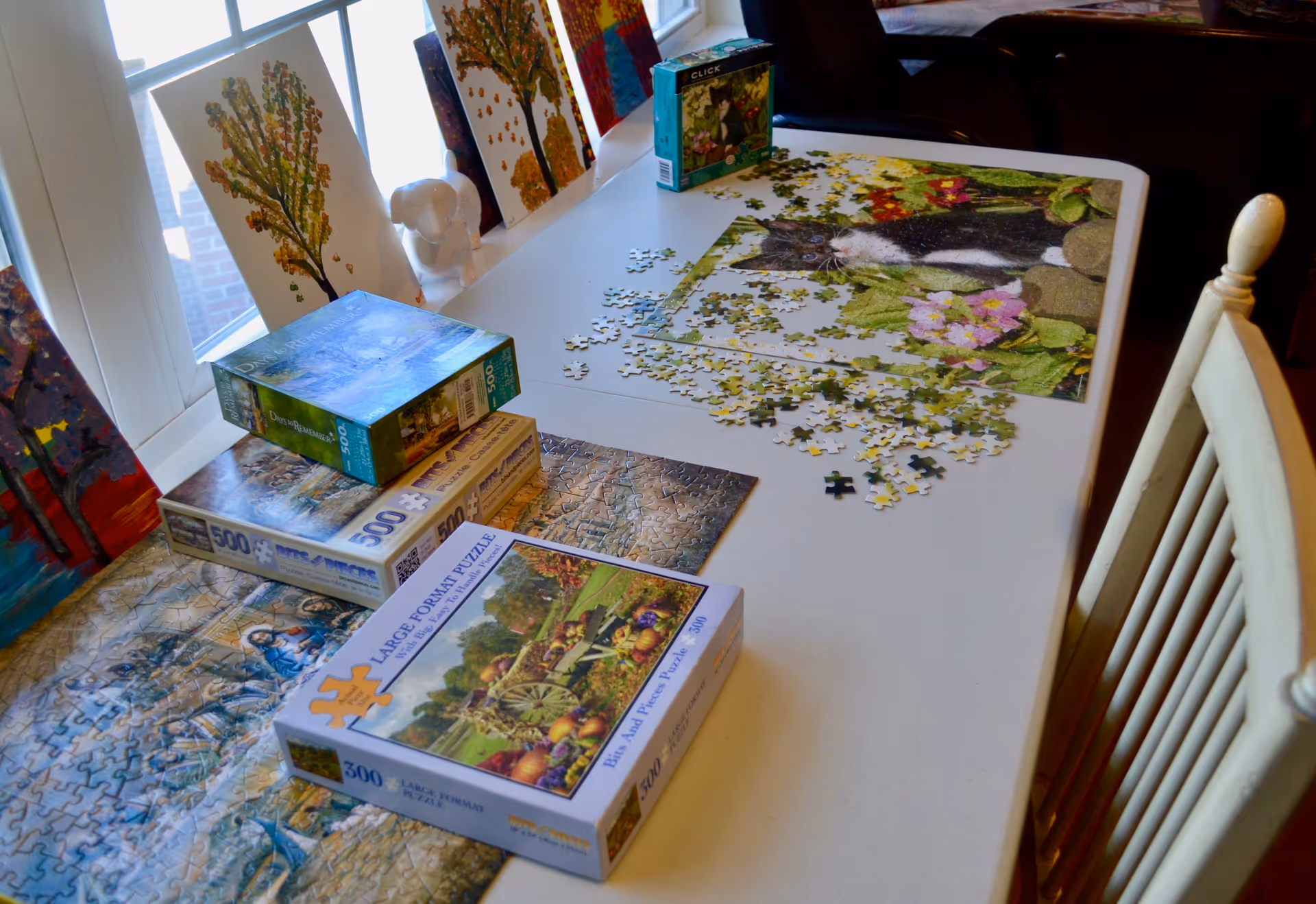 A white table near a window with several jigsaw puzzles in progress and puzzle boxes stacked on top. Completed and partially completed puzzles depict various scenes including a cat among flowers and a scenic landscape. Several colorful paintings of trees are propped up against the window sill.