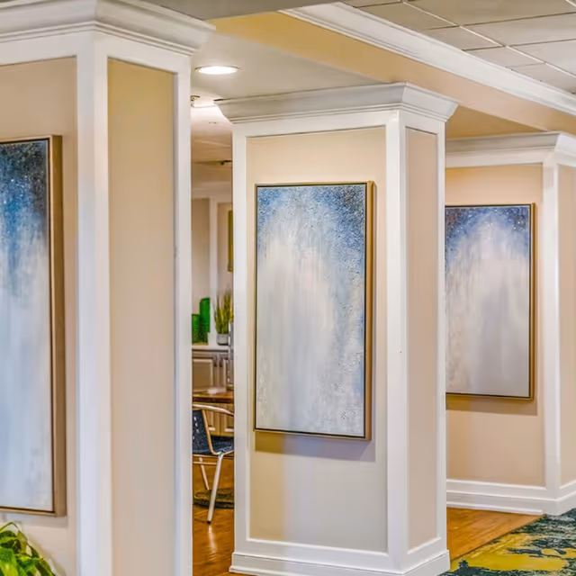 Interior view of a senior living facility hallway with beige walls and white trim. Two pillars display abstract paintings with blue and white tones. Part of a dining area with chairs and a table is visible in the background.