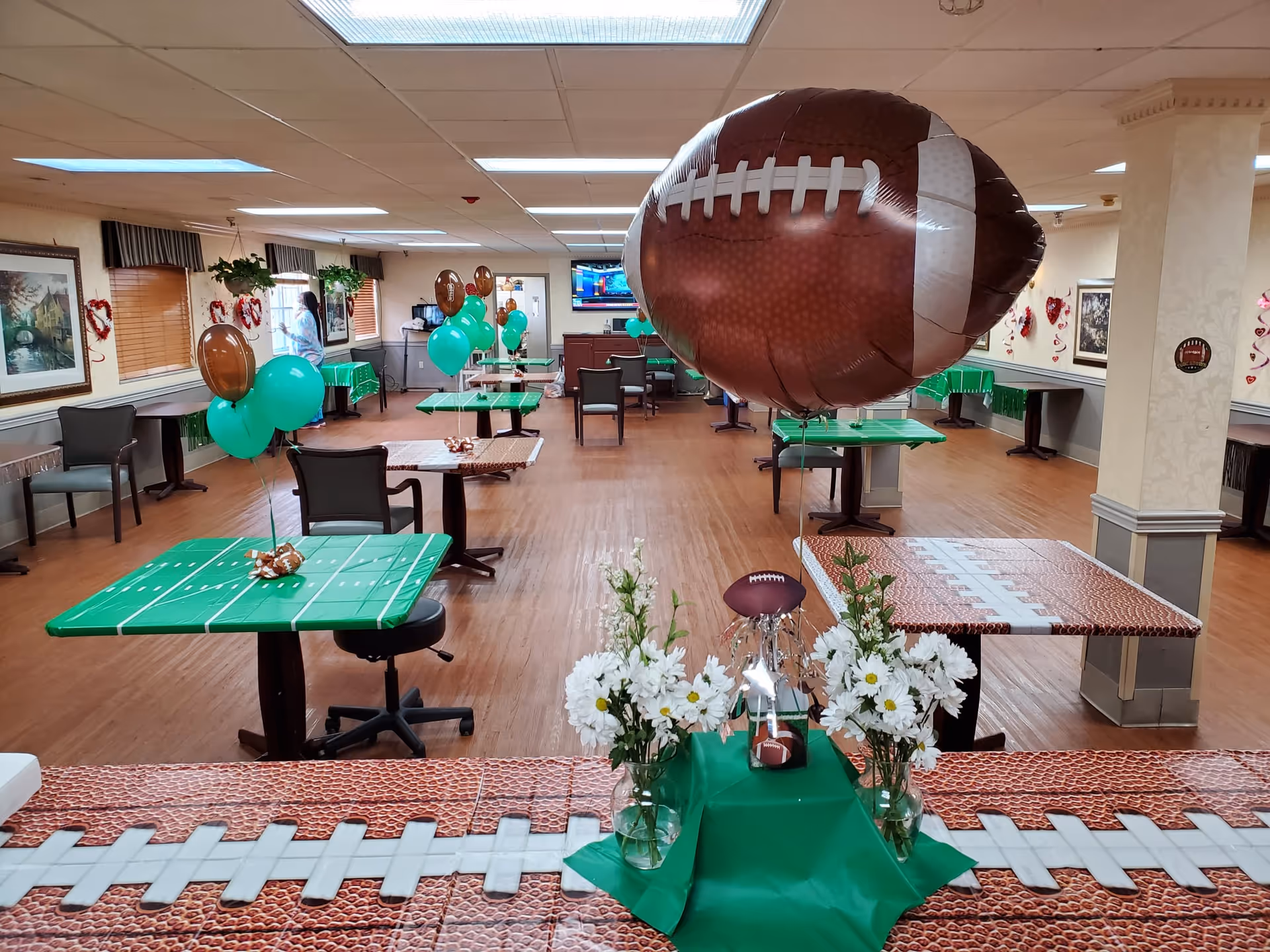 A decorated dining room with tables covered in football-themed tablecloths and balloons. The tables have green and brown football-themed decorations, including large football-shaped balloons and smaller balloons tied together. There are white flowers in vases on one of the tables. The room has wooden floors, beige walls, and several chairs around the tables. A television is mounted on the far wall.